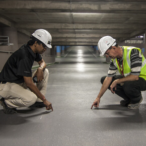 Two construction men with white hardhats pointing at the ground in a parking garage
