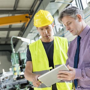 Two men, one in safety clothing and one in a dress shirt and tie looking and discussing information in a notebook