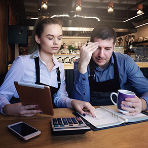 Man and woman in aprons sitting at table looking frustrated at calculator, accounting books