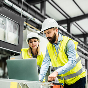 Two people in vests and hard hats looking over a laptop