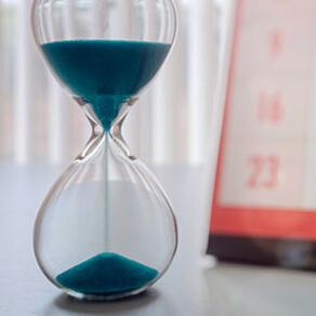 Picture of a clear hour glass filled with blue sand half way run through the glass and a calendar behind it
