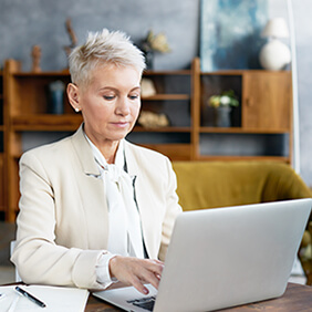 Beautiful trendy looking 50 year old businesswoman in stylish suit keyboarding on portable computer, sitting at office desk with smart phone and copybook. Technology and occupation concept