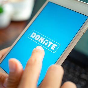 Close-up Finger pressing donate icon button on blue screen on white mobile phone with black laptop computer on wooden table. Donation online concept.