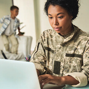 Woman in military uniform sitting in front of a laptop with another military member in the background