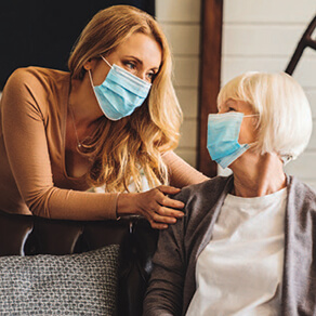 A younger woman with her hands on the shoulders of an older woman, both wearing medical face masks