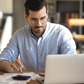 Man sitting looking at his laptop computer with papers and calculator and pen on table