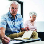 A older male and female sitting together while the male writes on a notepad with an agenda in his other hand and the female sits more in the distance typing on a computer.