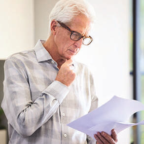 - Older man with glasses reading papers with hand on chin.