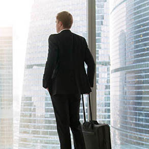 Young businessman with suitcase standing looking at full length window, dreaming to move only forward, high rental price, tenant looking for luxury home. Rear view. Business trip concept