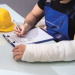 Person with a cast on filling out forms, hard hat on desk