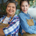 Senior mother with daughter, standing shoulder to shoulder wearing aprons