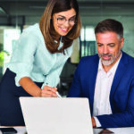 Man in a suit sitting at a desk looking at a computer while a woman standing to his right shoulder is pointing at the computer screen explaining what they are looking at