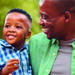 A close-up view of a 3-generation family, grandfather, father, and toddler, all with big smiles as the grandfather and father look inwards at the toddler.