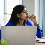 A woman sitting at a desk, with a computer and notepad in front of her, looking off to her left with a pondering expression on her face while biting her thumb nail.