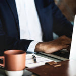 Torso of a man typing on a laptop with a cup and paper with pen on it