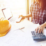 An engineer , architect woman using calculator for accounting and analyzing building structure from blueprint and paper chart in front of computer laptop at office work space.
