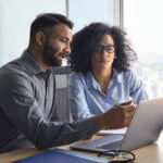 A man and woman sitting in front of a window looking at a laptop