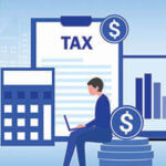 Cartoon like image of businessman sitting on a stack of coins working on a laptop with a calculator, clipboard with word "tax" on it and chart behind him