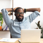 Man sitting at a desk with his hands behind his head smiling looking at his laptop