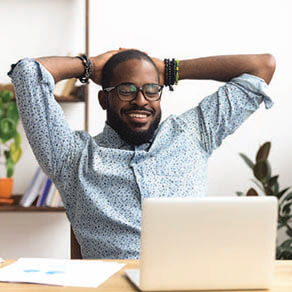 Man sitting at a desk with his hands behind his head smiling looking at his laptop