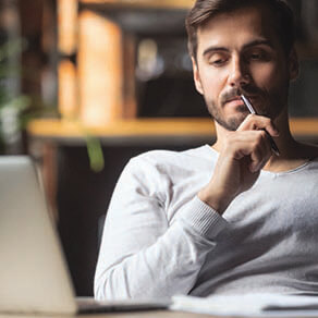 Man thinking with pen held up to his mouth looking at his laptop