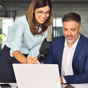 Man in a suit sitting at a desk looking at a computer while a woman standing to his right shoulder is pointing at the computer screen explaining what they are looking at